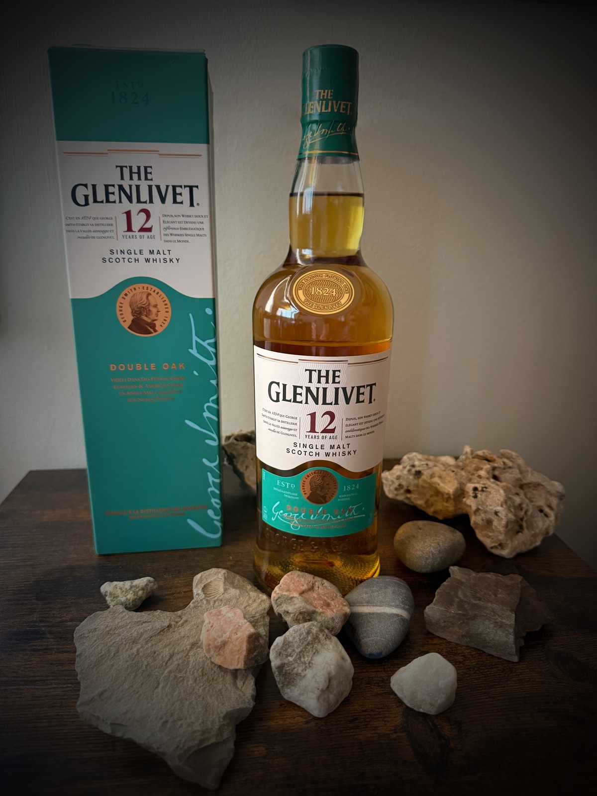 The Glenlivet 12 Year Old Double Oak bottle and its teal carton on dark wood, surrounded by pale limestone fragments and river pebbles